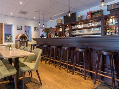 Sfeervolle hotelbar in Amrâth Hotel Alkmaar met houten bar, barkrukken, groene stoelen en warme verlichting.